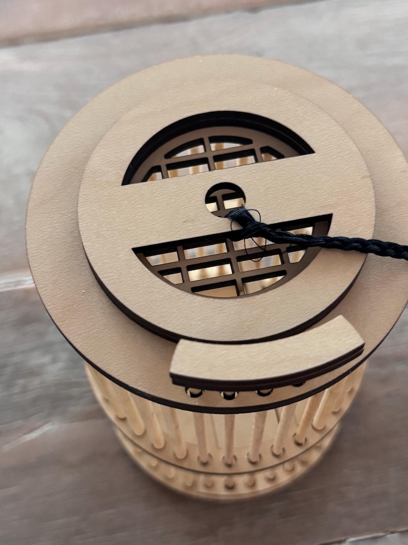 May include: A wooden lantern with a circular top featuring a decorative cutout and a black cord. The lantern's body is cylindrical, with vertical wooden supports. The design is natural and minimalist, suitable for home decor.