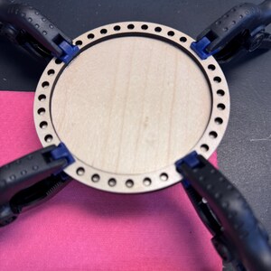 May include: A light-colored wooden craft circle with a series of small holes around the perimeter. The circle is held in place by four black clamps with blue tips, resting on a pink surface. Wooden dowels and a small ball are visible.
