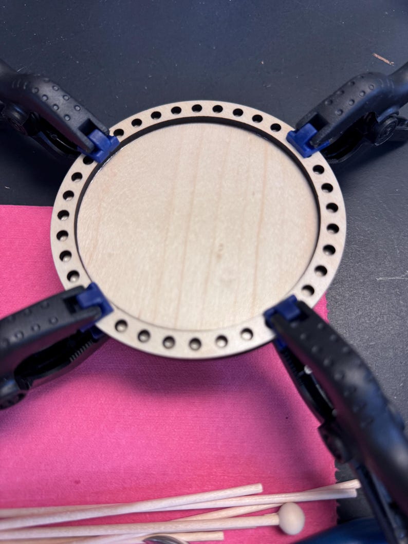 May include: A light-colored wooden craft circle with a series of small holes around the perimeter. The circle is held in place by four black clamps with blue tips, resting on a pink surface. Wooden dowels and a small ball are visible.