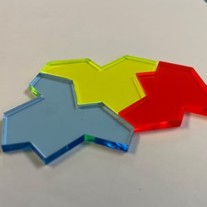 Op de afbeelding: Vier kleurrijke, doorschijnende spelstukken in blauw, geel en rood. Elk stuk heeft een unieke geometrische vorm met een gekerfd ontwerp. De stukken zijn gerangschikt op een wit oppervlak, waardoor een visueel aantrekkelijke opstelling ontstaat.