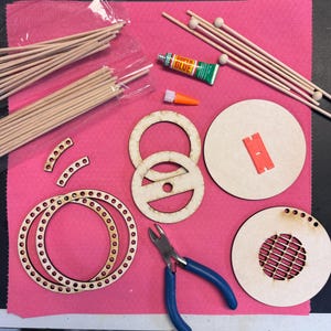 May include: A collection of craft supplies arranged on a pink surface. Includes wooden dowels, circular wooden pieces with holes and cutouts, small wooden balls, a tube of Super Glue, and blue-handled pliers. A small orange glue applicator is also present.
