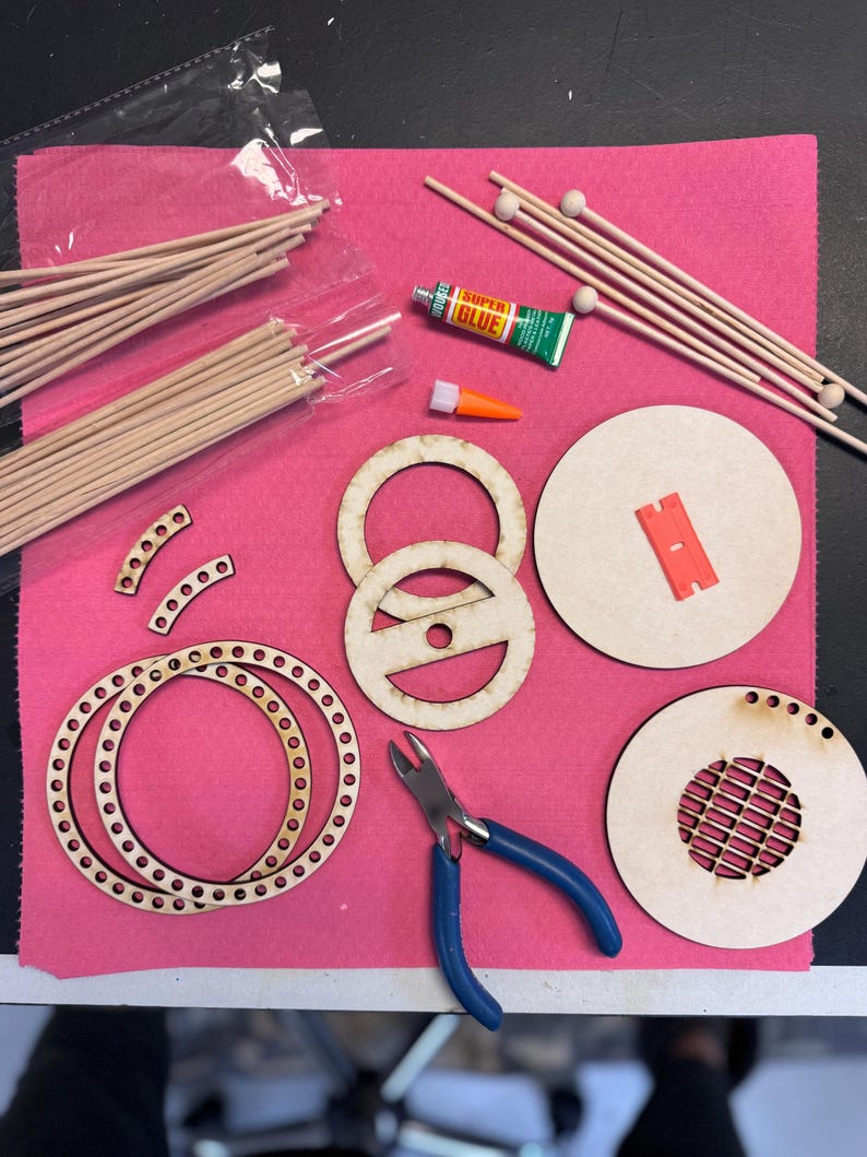 May include: A collection of craft supplies arranged on a pink surface. Includes wooden dowels, circular wooden pieces with holes and cutouts, small wooden balls, a tube of Super Glue, and blue-handled pliers. A small orange glue applicator is also present.