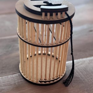 May include: A wooden cage-like container with a circular top and base. The cage is constructed of vertical wooden dowels held together by black bands. A black tassel hangs from the top. The container is made of light brown wood.