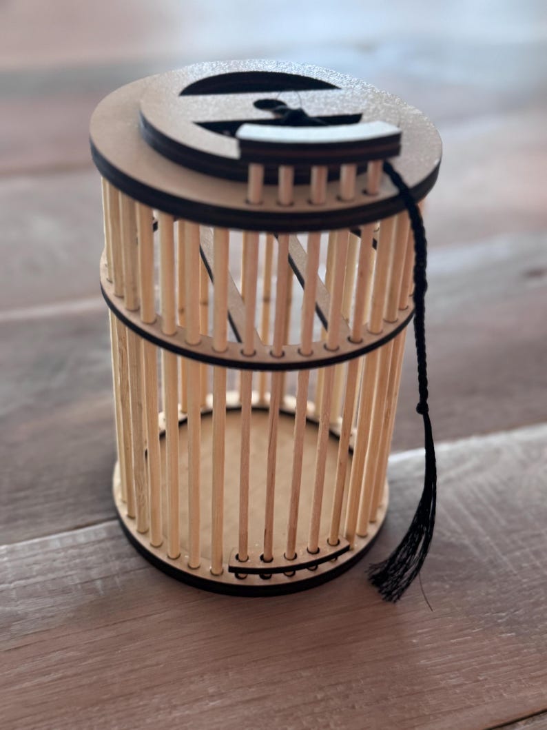 May include: A wooden cage-like container with a circular top and base. The cage is constructed of vertical wooden dowels held together by black bands. A black tassel hangs from the top. The container is made of light brown wood.