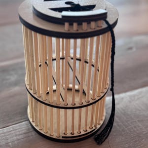 May include: A cylindrical wooden cage with vertical dowels and a circular top and bottom. The top has a decorative cutout and a black tassel attached. The cage is made of light-colored wood with dark brown accents.