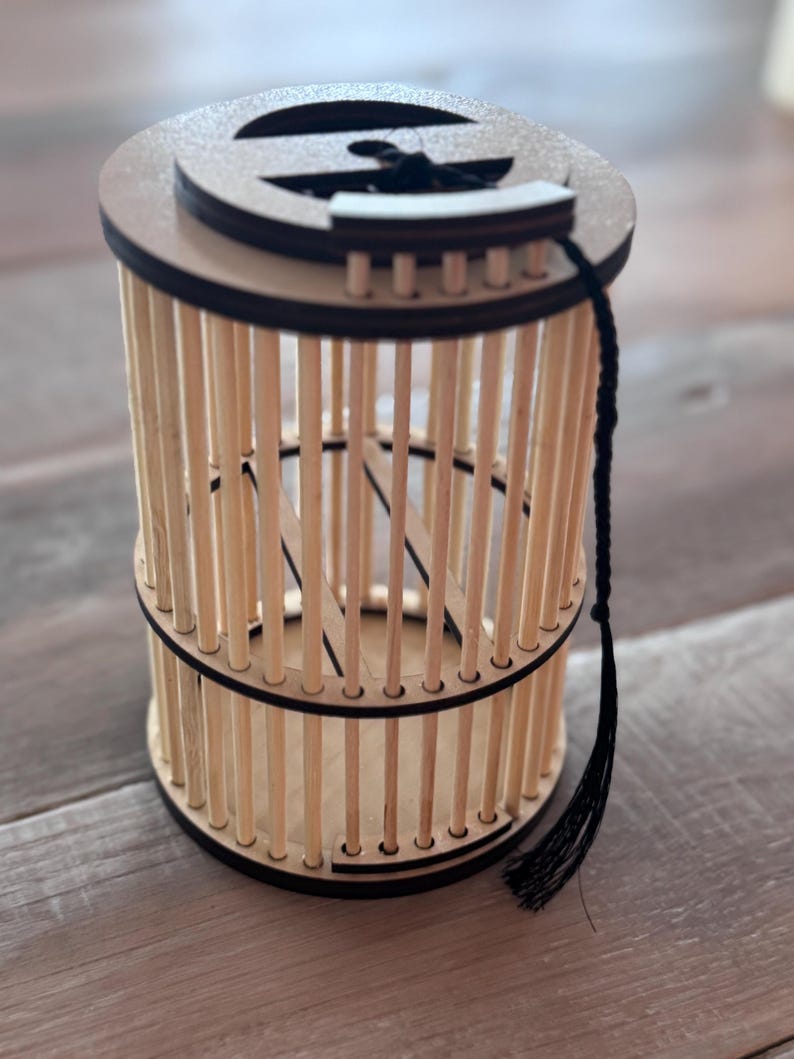 May include: A cylindrical wooden cage with vertical dowels and a circular top and bottom. The top has a decorative cutout and a black tassel attached. The cage is made of light-colored wood with dark brown accents.