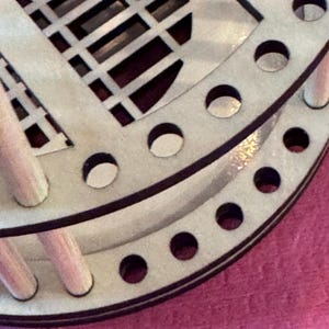 May include: Close-up of a light-colored wooden object with a circular design. The object has a series of small, evenly spaced holes around the edge and a geometric pattern cut into the surface. The wood has a natural, unfinished appearance.