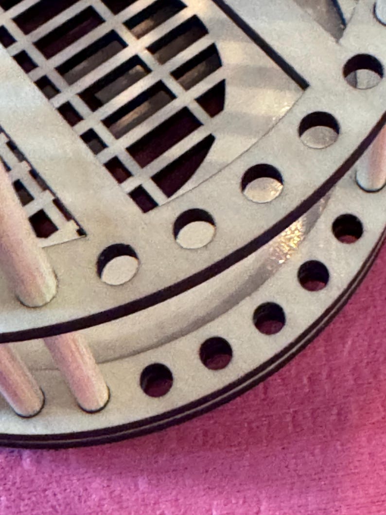 May include: Close-up of a light-colored wooden object with a circular design. The object has a series of small, evenly spaced holes around the edge and a geometric pattern cut into the surface. The wood has a natural, unfinished appearance.