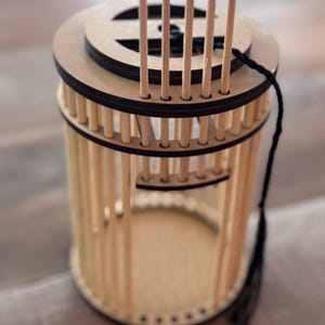 May include: A cylindrical wooden lantern with a circular top and base. The lantern features vertical wooden rods forming the sides, with a decorative top and a black tassel hanging from the side. The lantern is made of light-colored wood.