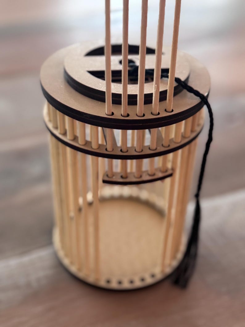 May include: A cylindrical wooden lantern with a circular top and base. The lantern features vertical wooden rods forming the sides, with a decorative top and a black tassel hanging from the side. The lantern is made of light-colored wood.