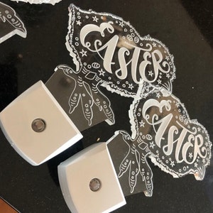 May include: Two white acrylic Easter night lights featuring a chick design and the word "Easter" in a decorative font. Each night light has a small, round, white light bulb.