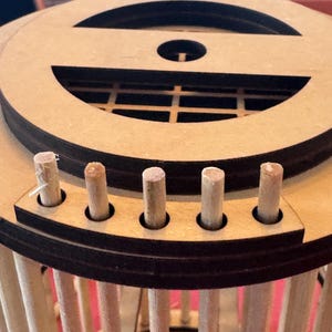 May include: A wooden craft project in progress, featuring a circular design with multiple layers and vertical dowels. The top layer has a decorative cutout, and small wooden pegs are inserted around the perimeter. The structure is unfinished.