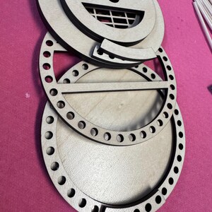 May include: A collection of unfinished wooden craft pieces, including circular frames with evenly spaced holes, and a decorative top with a grid pattern. The pieces are arranged on a pink surface, suggesting a crafting project or DIY kit.