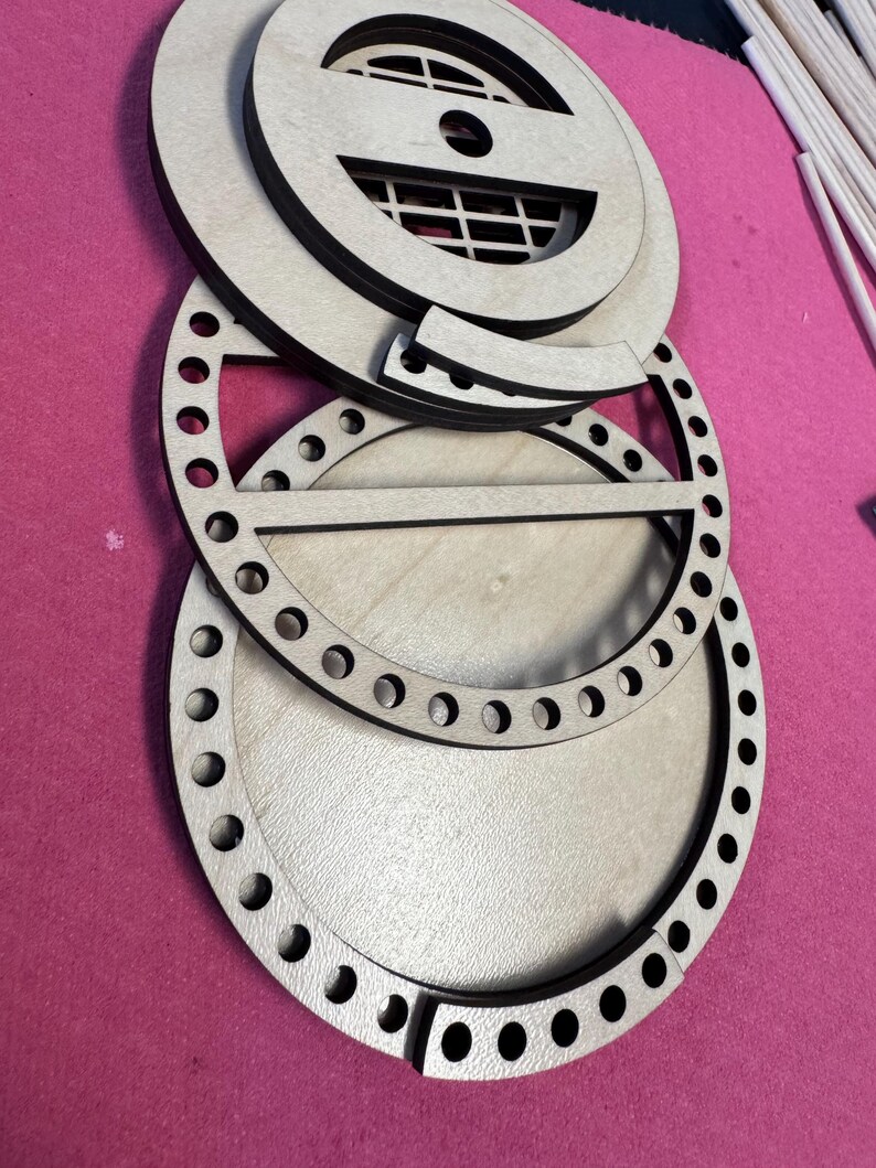 May include: A collection of unfinished wooden craft pieces, including circular frames with evenly spaced holes, and a decorative top with a grid pattern. The pieces are arranged on a pink surface, suggesting a crafting project or DIY kit.