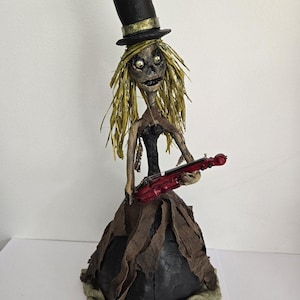 May include: A handcrafted folk art sculpture of a figure with a top hat and a red violin. The figure has a skeletal face, straw-like hair, and a tattered dress. The sculpture is made of paper mache and fabric.