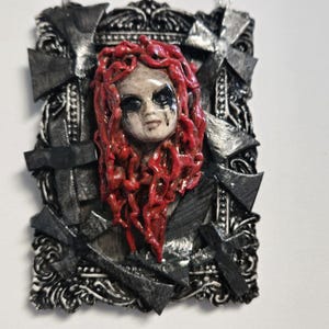 May include: A gothic pendant with a sculpted face, red hair, and dark eye makeup. The face is set within a black and silver frame with cross-shaped accents. The pendant has a textured, handcrafted appearance.