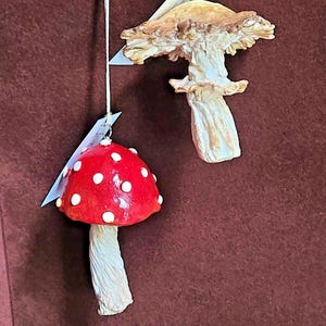 May include: Two hanging mushroom ornaments suspended by white strings. One ornament is a bright red mushroom cap with white spots and a textured cream-colored stem. The other is a realistic, textured brown mushroom with a wide, ruffled cap.