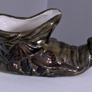May include: A ceramic planter shaped like a boot, with a dark green and brown glaze. The boot features a white interior and is decorated with relief mushrooms. The planter is designed for decorative use.