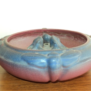 May include: A blue and pink ceramic planter with a decorative design of stylized butterflies and a central raised area with a hole. The planter has a crackled glaze finish.