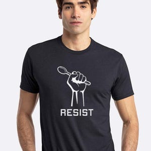 Puede incluir: Una camiseta gris oscuro jaspeada con un gráfico blanco de un puño sosteniendo una cuchara. El texto "RESIST" está impreso debajo del gráfico.
