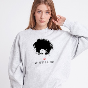 Op de afbeelding: Grijs sweatshirt met een zwart-witte illustratie van een persoon met de tekst "Why can't I be you?" gedrukt op de voorkant.