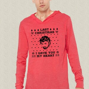 Op de afbeelding: Een rood langarm hoodie met een zwart-witte grafische print van een mannengezicht en de tekst "Last Christmas I Gave You My Heart".