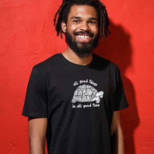 Könnte beinhalten: Ein Mann mit Dreadlocks trägt ein schwarzes T-Shirt mit einem weißen Motiv einer Schildkröte und dem Text "all good things in all good time".