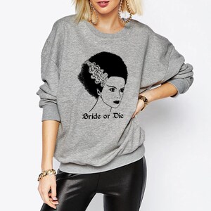 K&ouml;nnte beinhalten: Grauer Pullover mit einer schwarz-wei&szlig;en Illustration einer Frau mit einer Frisur, die an die Braut von Frankenstein erinnert. Der Text "Bride or Die" ist unter der Illustration gedruckt.