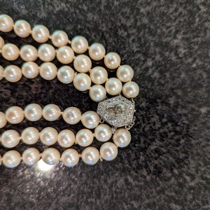 May include: A three-strand pearl necklace with a decorative silver clasp. The pearls are creamy white and evenly sized. The clasp is a geometric design with clear stones.