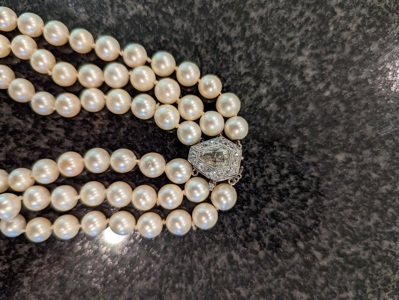 May include: A three-strand pearl necklace with a decorative silver clasp. The pearls are creamy white and evenly sized. The clasp is a geometric design with clear stones.
