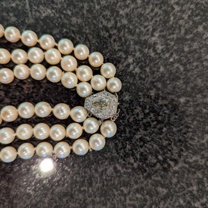 May include: A three-strand pearl necklace with a decorative clasp. The pearls are a creamy white colour and appear to be evenly sized. The clasp is silver-toned and features a geometric design with clear stones.