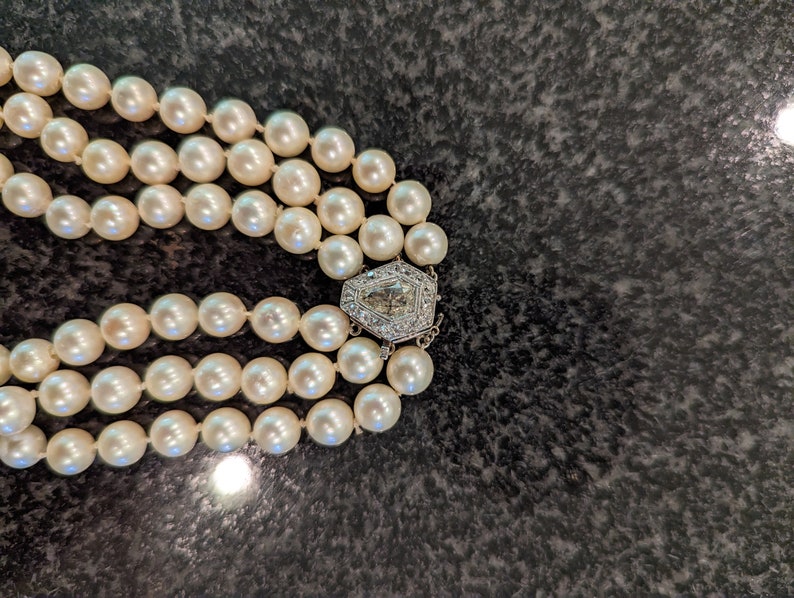 May include: A three-strand pearl necklace with a decorative clasp. The pearls are a creamy white colour and appear to be evenly sized. The clasp is silver-toned and features a geometric design with clear stones.