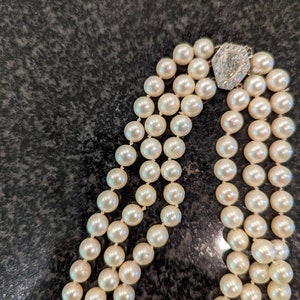 May include: A three-strand pearl necklace with a decorative clasp. The pearls are creamy white and evenly sized. The clasp is silver-toned with a central diamond-shaped accent.