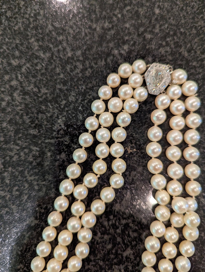 May include: A three-strand pearl necklace with a decorative clasp. The pearls are creamy white and evenly sized. The clasp is silver-toned with a central diamond-shaped accent.