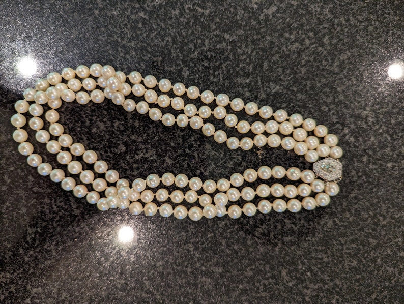 May include: A triple-strand pearl necklace with a decorative clasp. The pearls are a creamy white hue and appear uniform in size. The clasp is silver-toned and features small, clear stones.