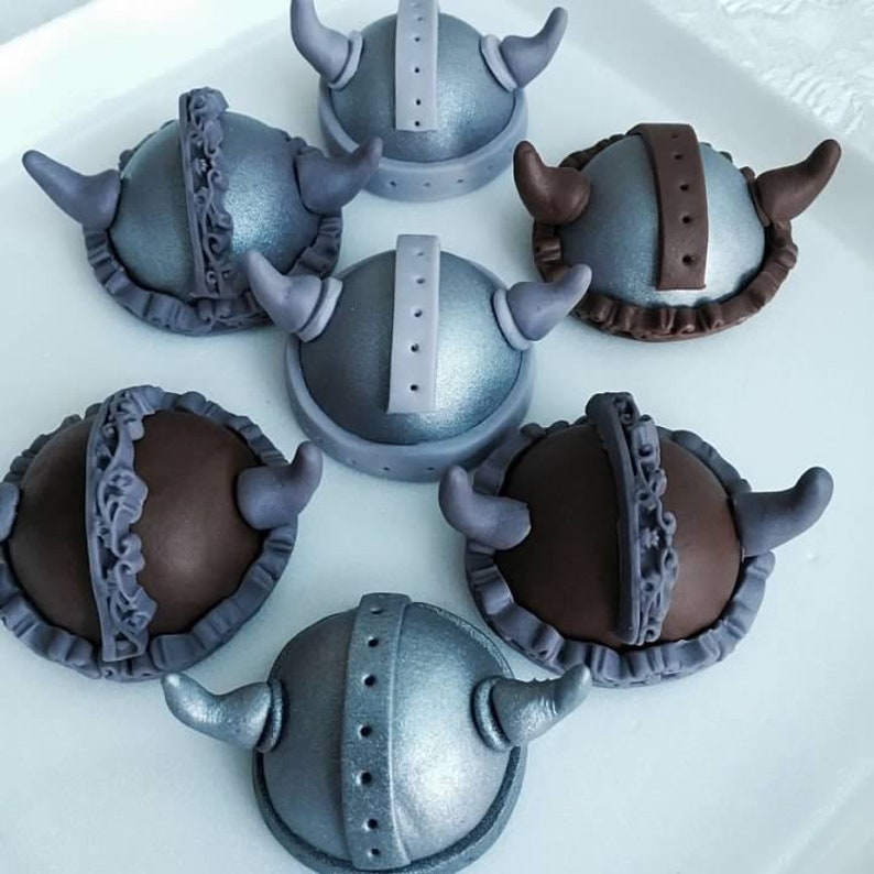 May include: Six miniature Viking helmet cupcakes decorated with silver and brown fondant. The helmets have horns and intricate details.