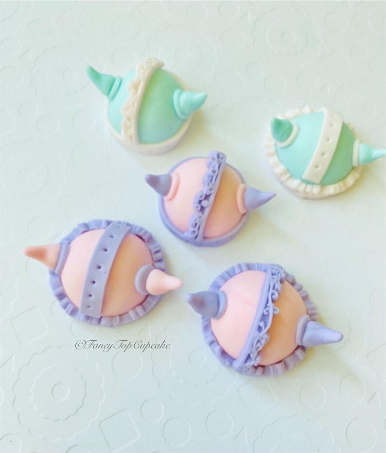May include: Five pastel-colored fondant cupcake toppers shaped like hats with horns. The toppers are decorated with white lace and purple or blue accents. The toppers are on a white background. @FancyTopCupcake
