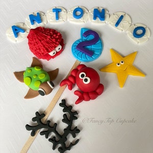 May include: A set of colorful fondant cake toppers featuring a red crab, a green turtle, a yellow starfish, a blue seashell with a purple number 2, a red and white sea anemone, and white sand dollar letters spelling out "ANTONIO".