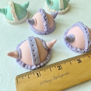 May include: Five fondant cupcake toppers shaped like helmets with horns. The helmets are in shades of pink, purple, and light blue with white accents. The toppers are approximately 1 inch in diameter.