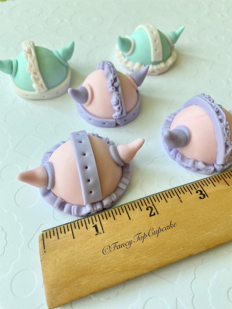 May include: Five fondant cupcake toppers shaped like helmets with horns. The helmets are in shades of pink, purple, and light blue with white accents. The toppers are approximately 1 inch in diameter.