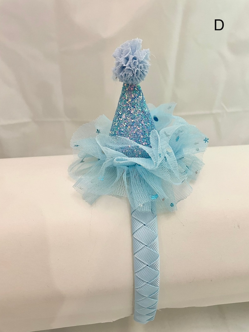 May include: A light blue headband with a blue tulle bow and a sparkly blue party hat on top. The headband is made of a braided fabric.