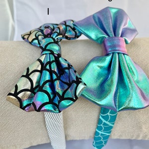 May include: Two mermaid-themed headbands with iridescent bows. One headband has a teal and gold scale pattern, the other has a solid teal scale pattern.
