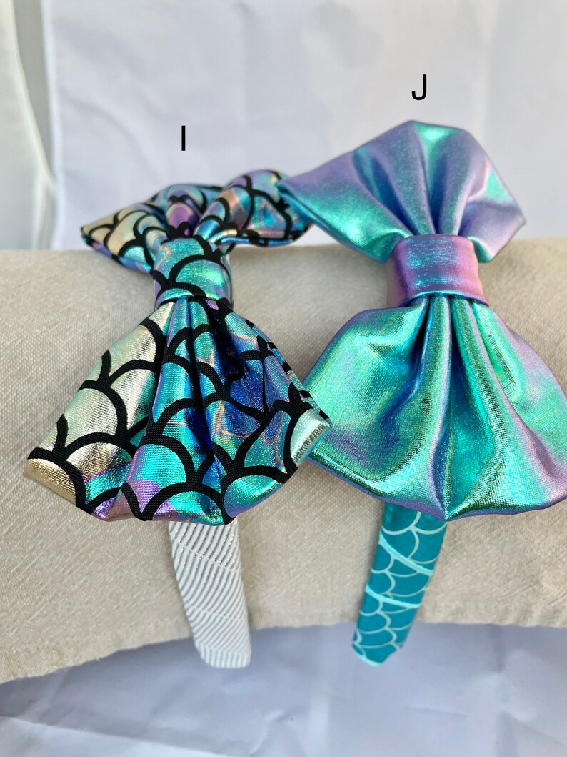 May include: Two mermaid-themed headbands with iridescent bows. One headband has a teal and gold scale pattern, the other has a solid teal scale pattern.