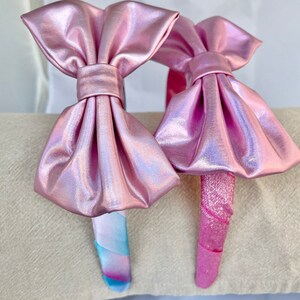 May include: Two pink iridescent headbands with large bows. One headband has a pink and blue swirl pattern and the other has a solid pink pattern.