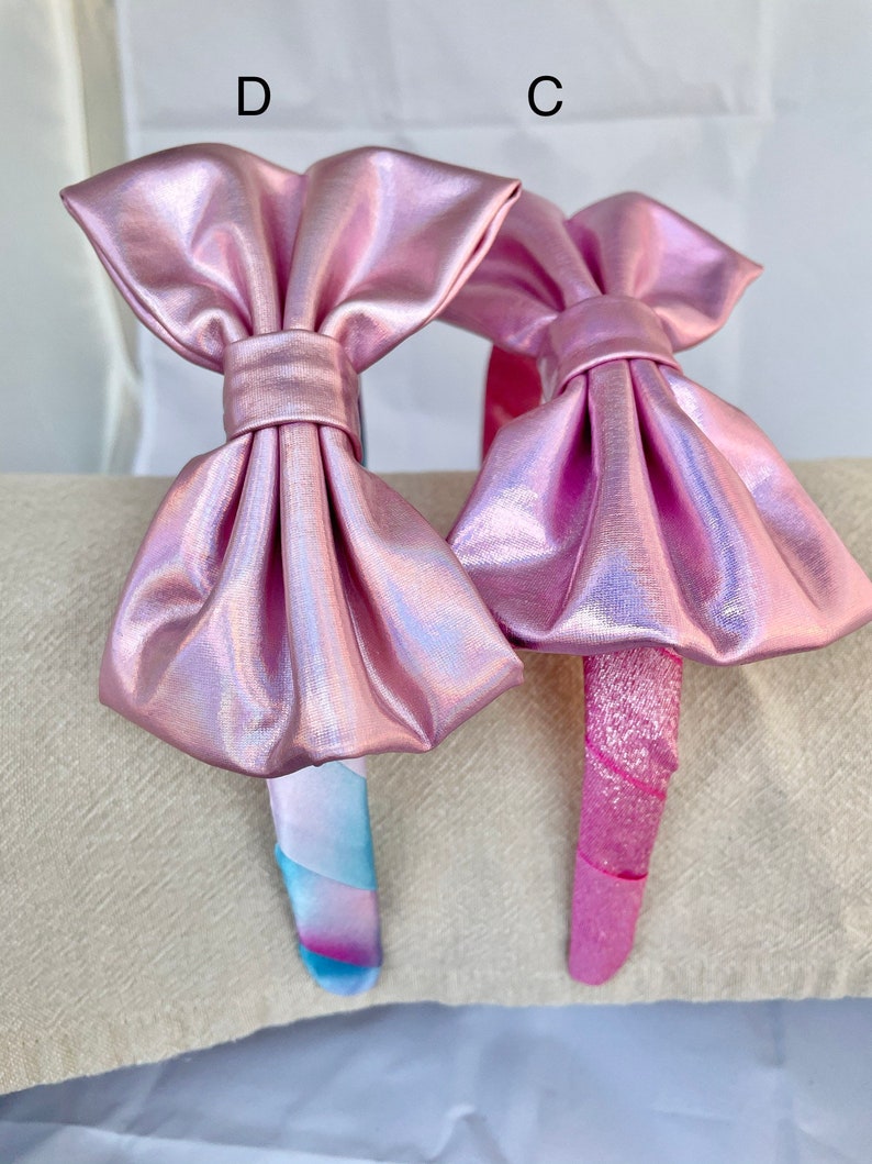 May include: Two pink iridescent headbands with large bows. One headband has a pink and blue swirl pattern and the other has a solid pink pattern.