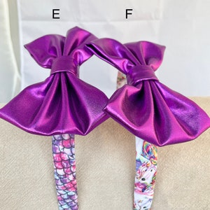 May include: Two purple metallic bow headbands with different patterned bands. One headband has a mermaid scale pattern and the other has a unicorn pattern.