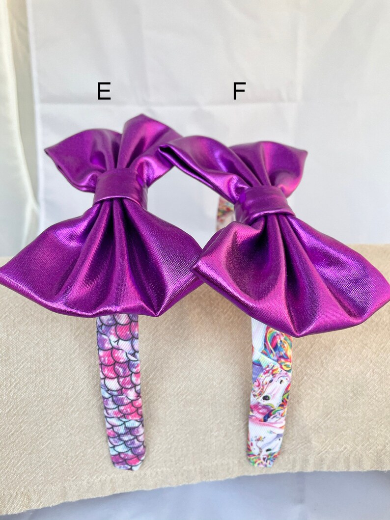 May include: Two purple metallic bow headbands with different patterned bands. One headband has a mermaid scale pattern and the other has a unicorn pattern.