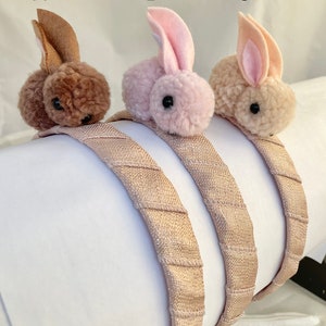 May include: Three headband hair accessories with bunny pom-poms. The headbands are a light brown color with a textured fabric. The bunny pom-poms are brown, pink, and beige.