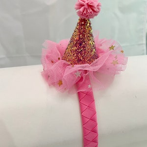 May include: Pink headband with a pink and gold glitter party hat with a pink pom pom on top. The hat is surrounded by a pink tulle with gold stars.
