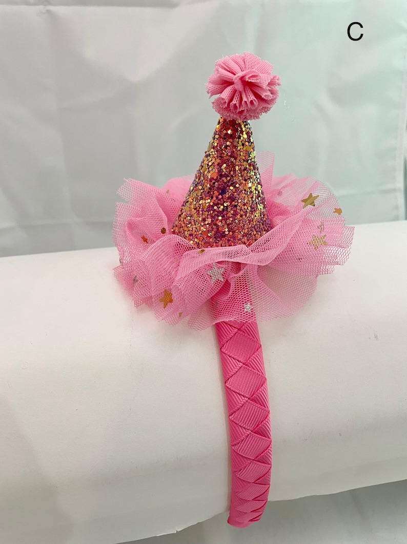 May include: Pink headband with a pink and gold glitter party hat with a pink pom pom on top. The hat is surrounded by a pink tulle with gold stars.